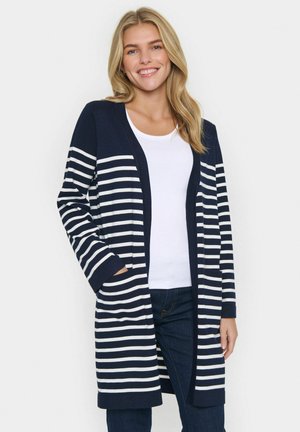Glunderende vrouw in een marineblauwe en witte gestreepte lange cardigan over een witte top en donkere jeans, staande tegen een effen achtergrond.