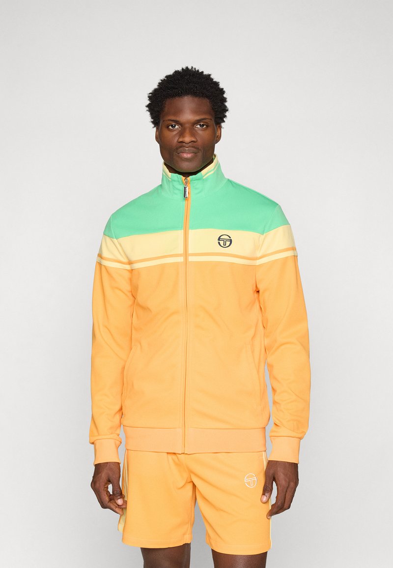 Oransje og teal joggedress med høy krage. Har horisontale striper i gul og hvit. Glidelås foran og logo på brystet.