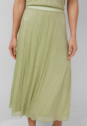 Vrouw draagt een lichtgroene geplooide midi-rok met subtiele glans, handen ontspannen langs het lichaam, staand tegen een effen grijze achtergrond.