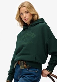 Sudadera verde con un gran bolsillo frontal y capucha ajustable. Presenta el texto 'Superdry NYC' en un verde más claro, llevada sobre una camisa de cuadros.