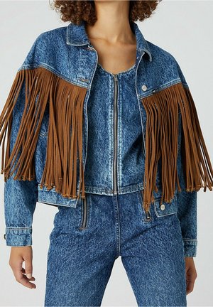 Kobieta w niebieskiej kurtce jeansowej z efektem acid wash z brązowymi frędzlami na ramionach, dopasowanym topie jeansowym na zamek i spodniach.