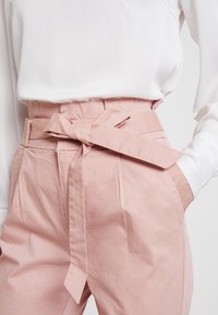Pantalons taille haute de couleur rose clair avec une ceinture à nouer, dotés de plis, de poches latérales et d'un tissu texturé. Associés à un chemisier blanc.