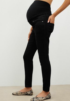 Zwangere persoon die zwarte zwangerschapsjeans met een rekbare tailleband en platte schoenen met slangenvormig patroon draagt, staand opzij.