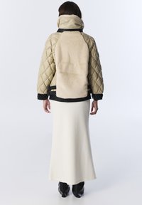 Femme portant une veste matelassée beige avec un grand panneau en fourrure au dos, une longue jupe crème et des bottes noires, se tenant devant un fond blanc.