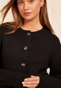 Black knitted cardigan featuring a round neckline, long sleeves, five buttons down the front, and ribbed cuffs and hem.