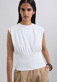 Haut blanc sans manches avec une taille froncée, un col rond et une texture lisse, porté avec un pantalon beige clair et un bracelet large foncé.