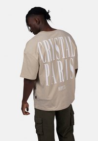 Beżowa oversize koszulka z białym napisem "CRYSTAL PARIS" na plecach i "DEPT.75" poniżej, wykonana z miękkiej bawełny.