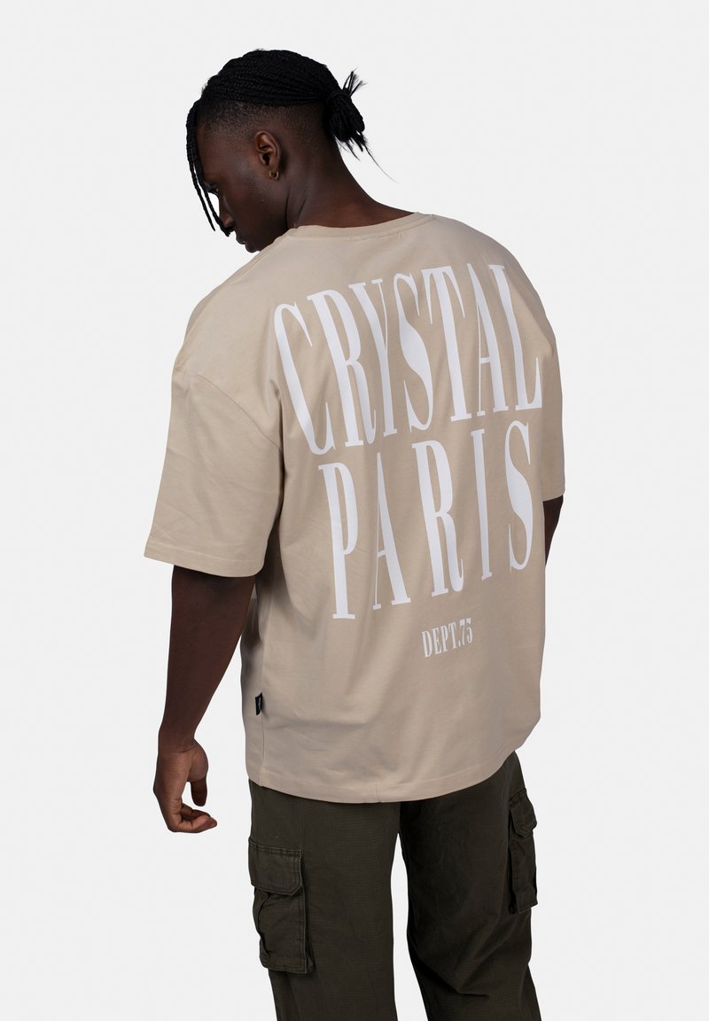 Beżowa oversize koszulka z białym napisem "CRYSTAL PARIS" na plecach i "DEPT.75" poniżej, wykonana z miękkiej bawełny.