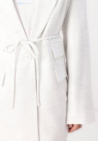 Giacca bianca con texture, cintura legata, chiusura con grandi bottoni e due tasche a toppa, caratterizzata da un sottile motivo a griglia e maniche a campana.