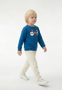 Sweat-shirt bleu avec broderies sur le thème de Noël, associé à un pantalon en velours côtelé crème et des baskets blanches. Enfant marchant sur un fond clair.