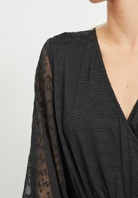Svart klänning med ett texturerat mönster, genomskinliga ärmar och djup V-ringning, som framhäver ett lätt material och avslappnad passform.