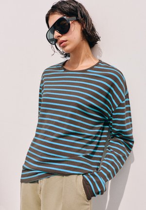 Persoon draagt een oversized zwarte zonnebril, een bruinblauw gestreept shirt met lange mouwen en beige broek, leunend tegen een effen lichte muur.