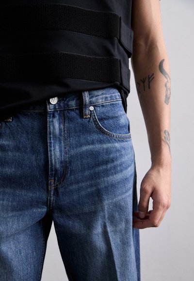 Fakó kék farmereken kontrasztos cérna, öt zsebes stílus, fém gombos záródás és slim fit fazon található. Fekete ujjatlan top, textúrázott pánttal.