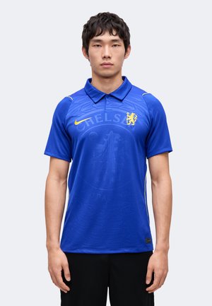 Man draagt blauw Chelsea voetbalshirt met geel Nike-logo en clubwapen, staand tegen een eenvoudige lichte achtergrond.