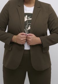 Blazer marron sur mesure porté sur un t-shirt blanc avec un motif floral vert; tissu lisse avec épaules structurées et poches.