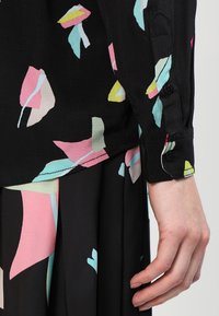 Blusa negra con patrones abstractos coloridos en rosa, azul claro y amarillo. Cuenta con puños abotonados y una tela lisa y ligera.