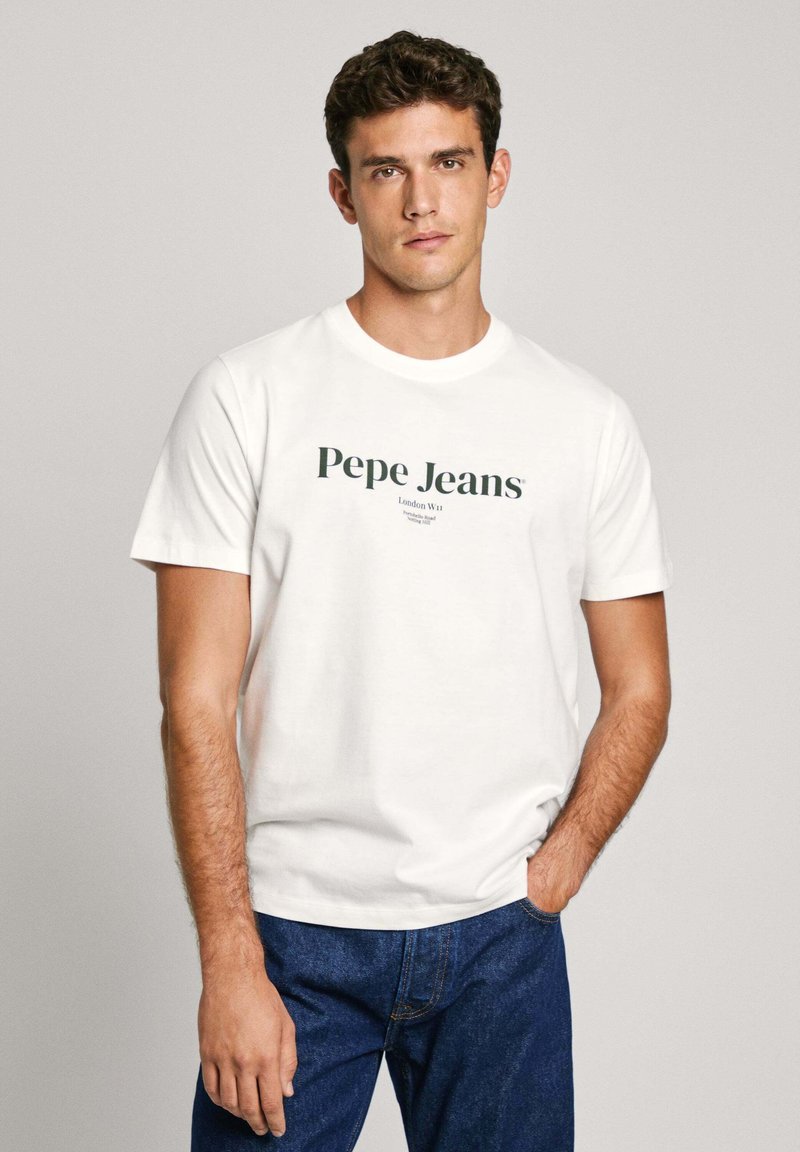 Camiseta blanca de algodón con mangas cortas que presenta el logo y el texto "Pepe Jeans" en verde. Combinada con unos jeans de mezclilla azul oscuro. Diseño sencillo.