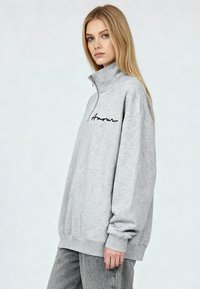Sweat-shirt gris zippé avec un col montant et le texte noir « Amour » sur le devant. Il a une coupe décontractée avec des poignets et un ourlet côtelés.