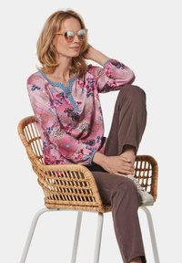 Rosa gemusterte Bluse mit blauen Akzenten, floralen Motiven und langen Ärmeln. Getragen mit braunen Hosen und weißen Schuhen, sitzt in einem Korbstuhl.