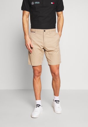 Mann trägt beige Shorts, weiße Sneakers und ein schwarzes Poloshirt mit Logos auf einfachem hellem Hintergrund.