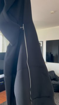 Melnā zip-up kapuce ar augstu apkakli, strukturētu audumu un redzamu baltu rāvējslēdzi gar malām, izcelta iekštelpu vidē.