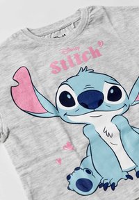 Lichtgrijs shirt met een grote kleurrijke cartoon Stitch-karakter, roze harten en de tekst "Disney Stitch" in het roze erboven.