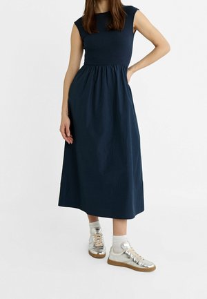 Kobieta w granatowej, bezrękawnikowej sukience midi z marszczonym pasem, w białych skarpetkach i metalicznych srebrnych sneakersach, stojąca na białym tle.