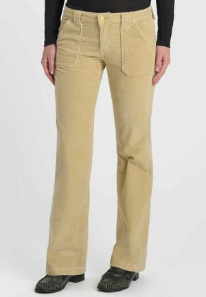 Beige corduroy broek met wijde pijpen, voorzakken, gedragen door een persoon die staat, gecombineerd met zwarte schoenen en een zwarte top met lange mouwen.