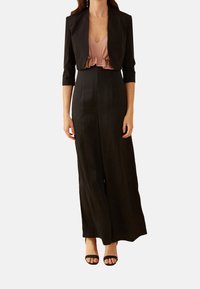 Černý overal s upnutým vrškem a širokými nohavicemi, pod kterým je černý zkrácený blazer. Hladká textura a minimální detaily; doplněno černými sandály.