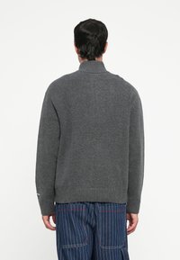 Donkergrijs sweater met een hoge kraag, geribbelde onderkant en een losse pasvorm. Gebreide textuur zonder zichtbare patronen. Achteraanzicht toont eenvoud in het ontwerp.