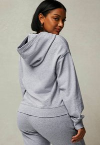 Grauer Kapuzenpullover aus weichem Material mit großer Kapuze, gerippten Bündchen und Saum. Das Modell wird von hinten gezeigt und präsentiert einen lässigen Schnitt.