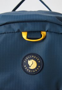 Sötétkék Fjällräven hátizsák sárga hurkolt fogantyúval és kerek logóval, amelyen egy róka feje és a "Fjällräven Bergtagen" felirat látható.