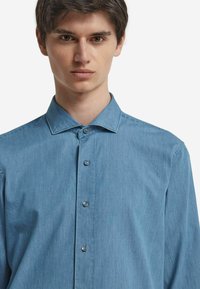 Ljust blå denimknapp-up skjorta med spetsig krage, långa ärmar och en slät textur. Har fem framknappar och diskret sömnad.