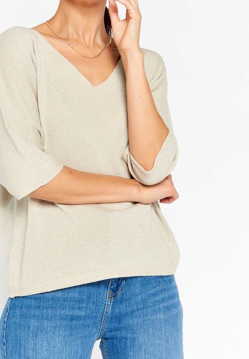 Femme portant un haut en maille beige à col en V avec des manches trois-quarts et un jean bleu, main près du visage et bras croisé sur la taille.