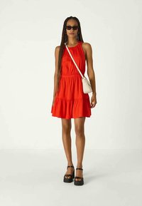Vestido vermelho sem mangas com um design em camadas, cintura ajustada e uma bainha ampla. Combinado com sandálias pretas de plataforma e uma pequena bolsa branca a tiracolo.