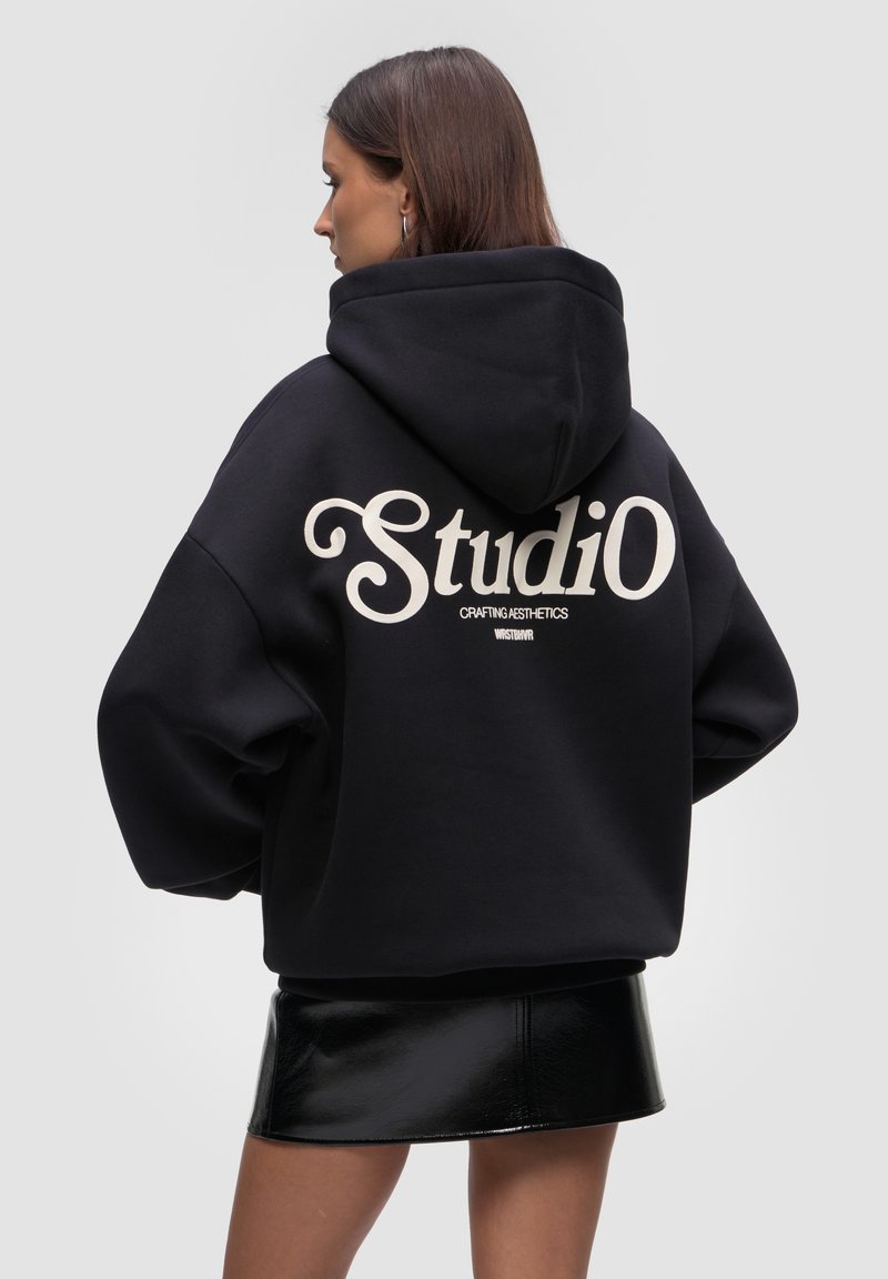 Czarna bluza z kapturem o oversize'owym kroju, z dużym białym napisem "Studio" na plecach. Miękka w dotyku, z kapturem na sznurku i ściągaczami przy mankietach.