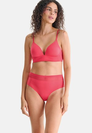 Femme aux cheveux bouclés portant un soutien-gorge corail assorti et une culotte taille haute, debout devant un fond blanc uni.