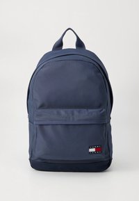 Mochila de nylon azul-marinho com um bolso frontal com fecho, alças ajustáveis e uma pega superior. Apresenta um emblema de logo com detalhes em vermelho, branco e azul.