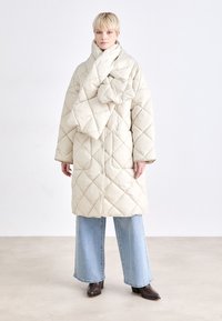 Casaco puffer beje acolchoado com grandes bolsos de remendo e uma gola alta, combinado com jeans azuis de perna larga e botins castanhos.