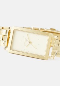 Orologio rettangolare d'oro con un design minimalista, che presenta un quadrante giallo chiaro, indicatori delle ore sottili e un bracciale in metallo a maglie.