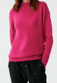Fuchsia gestrickter Pullover mit hohem Kragen, langen Ärmeln und geripptem Saum. Weiche Textur, lockere Passform, kombiniert mit schwarzen Hosen.