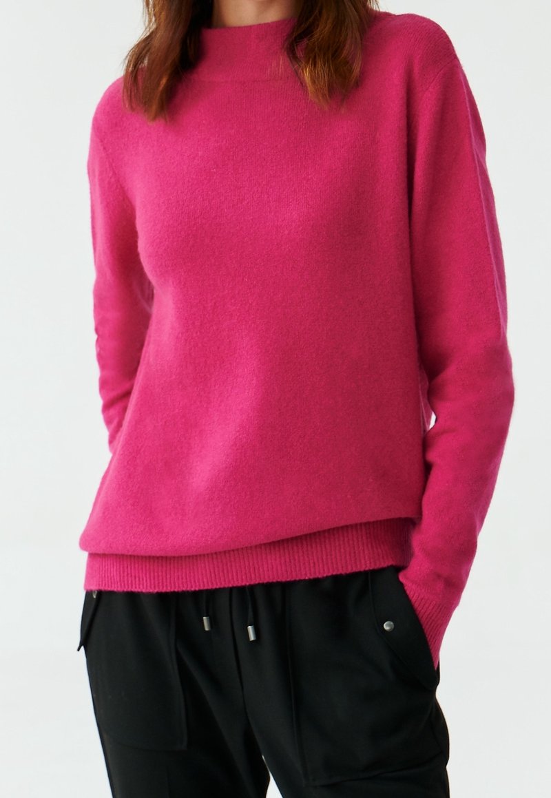 Fuchsia gestrickter Pullover mit hohem Kragen, langen Ärmeln und geripptem Saum. Weiche Textur, lockere Passform, kombiniert mit schwarzen Hosen.
