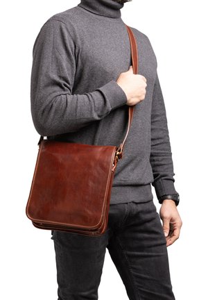 Man draagt een grijze coltrui en zwarte broek en draagt een bruine leren schoudertas met gestikte randen.