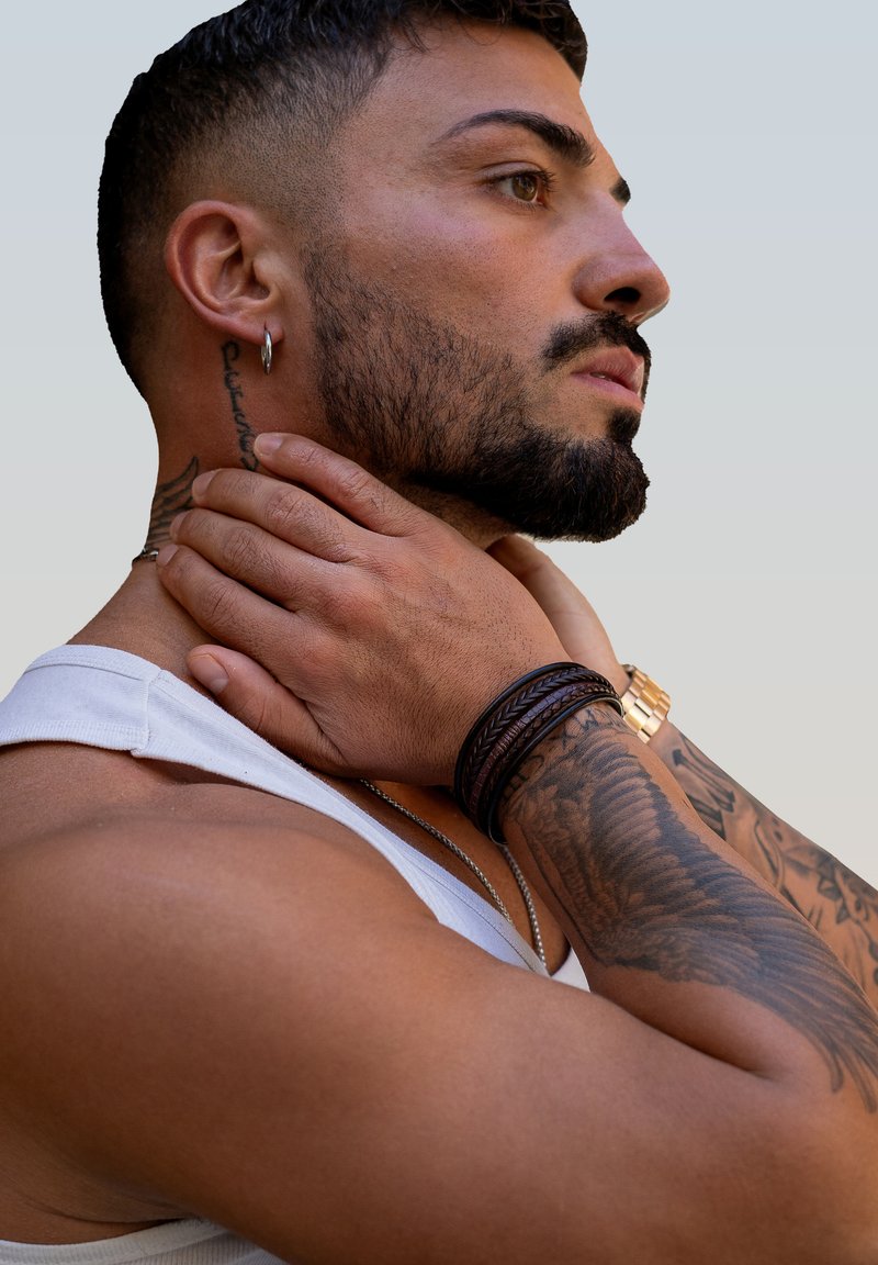 Close-up of a person wearing a white tank top, featuring dark braided leather bracelets, metallic watch, and visible tattoos on tan skin.