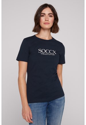 T-shirt en coton marine à manches courtes, col rond, avec le texte blanc "SOCCX TIMEELESS EDITION" sur la poitrine, associé à un jean en denim.