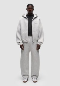 Felpa grigia chiara con cappuccio e tasche frontali, abbinata a pantaloni della tuta larghi coordinati. Una maglietta nera e sneakers bianche completano l'outfit.