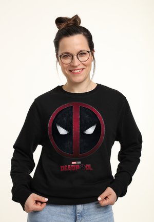 Uśmiechnięta kobieta w okularach i z włosami upiętymi w koka, ubrana w czarną bluzę Deadpool i jasnoniebieskie dżinsy, na jednolitym tle.