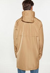 Smėlinis vandeniui atsparus džemperis su kapišonu, pasižymintis lygiu paviršiumi, nugaros ventiliacija ir ilgomis rankovėmis. Jis turi tiesų kirpimą ir minimalistinį dizainą.