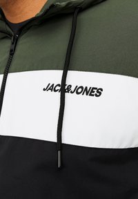 Kaputna jakna u maslinasto zelenoj, bijeloj i crnoj boji. Ima odvažne logotipe "JACK & JONES", debele špage i dijelove različitih boja.
