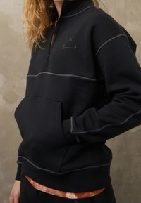 Sudadera negra con cremallera, acentos y costuras grises, con un bolsillo frontal y cuello alto. El material parece suave y texturizado.